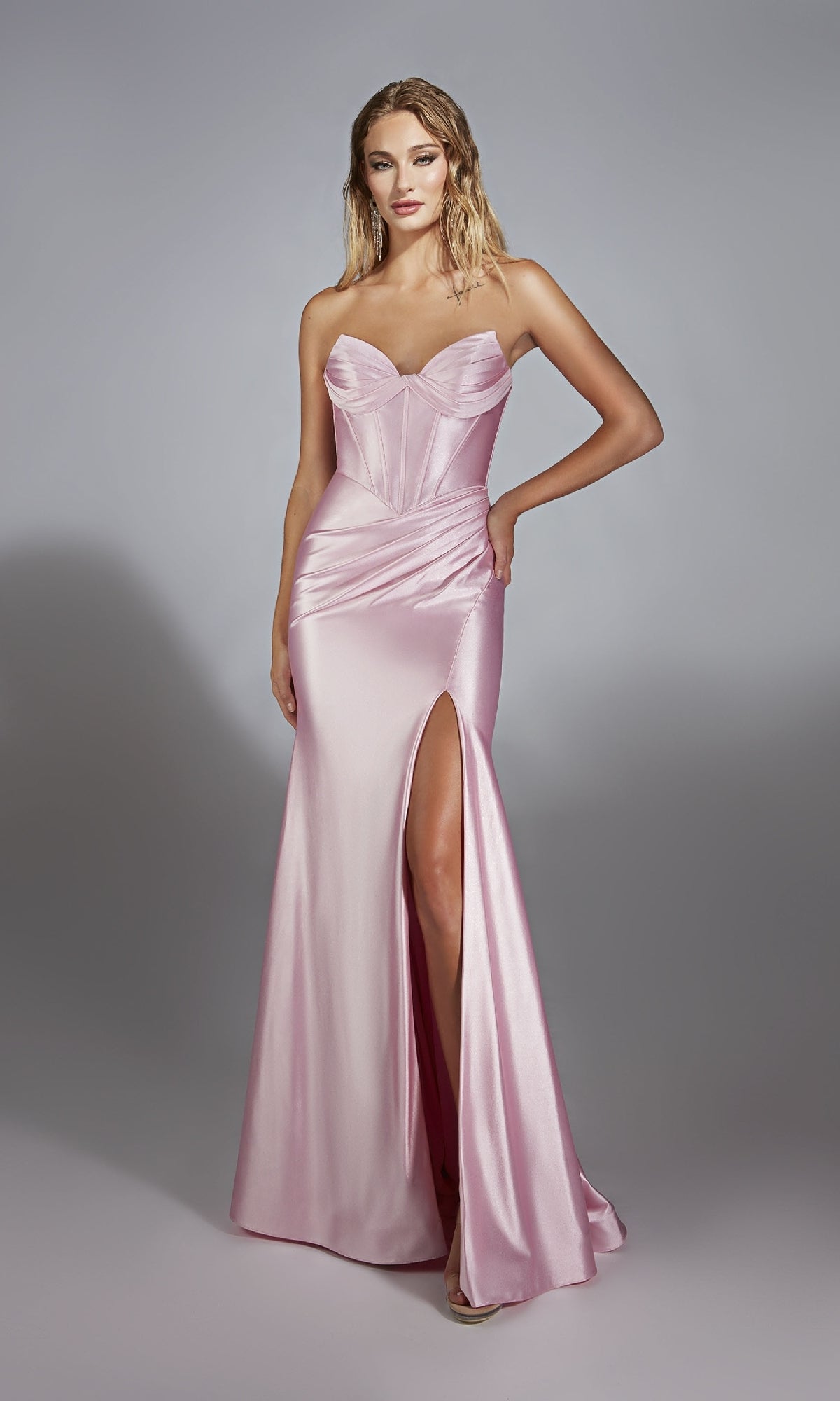 A woman wears the Alyce 61733 Strapless Long Corset Prom Dress in light pink satin with a sweetheart neckline and thigh-high slit, standing against a plain gray background.